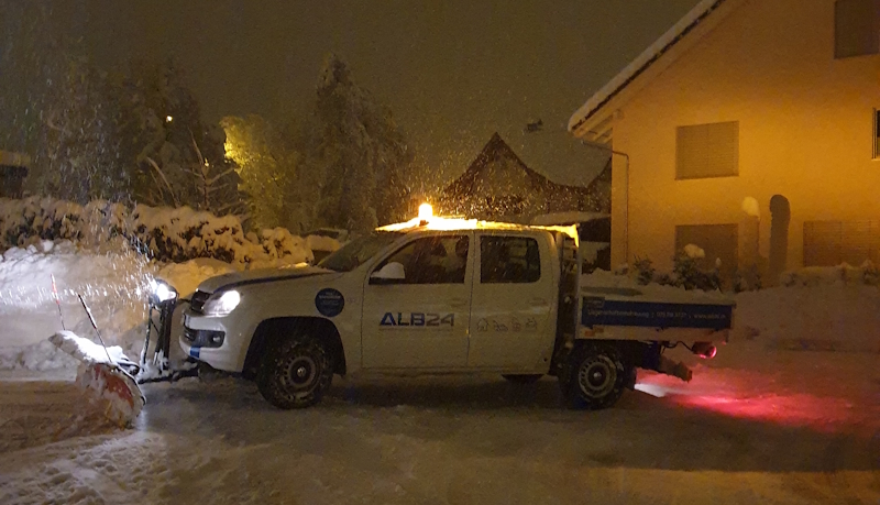 Fahrzeug der ALB24 GmbH mit Schneepflug räumt nachts bei Schneefall eine Zufahrt